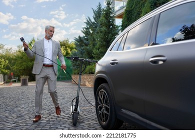 Businessman in suit on way to work with eletric scooter and charging his electric car. Concept of eco commuting and green transportation.