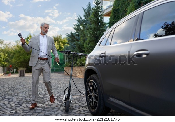 Businessman in suit on way to work with\
eletric scooter and charging his electric car. Concept of eco\
commuting and green\
transportation.
