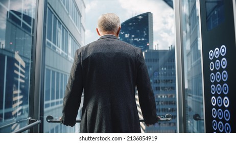 Businessman in a Suit Riding Glass Elevator to Office in Modern Business Center. Middle Aged Male Looking at Modern Downtown Skyscrapers Out of the Panorama Window in the Lift. Back Turned to Camera. - Powered by Shutterstock