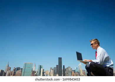 Businessman In A Suit Sitting Outdoors Using Laptop Computer Above The City Skyline Under Clear Blue Sky Copy Space