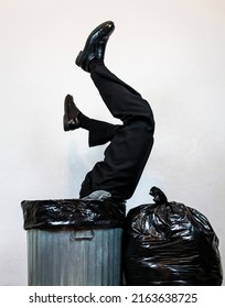 Businessman in Suit Stuck Upside Down in Metal Trash Can Next to Garbage Bag Pile. Concept of Over a Barrel. Thrown Away by Capitalism and Greed.