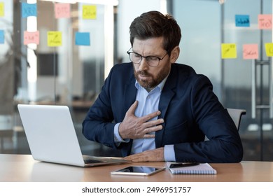 Businessman in suit suffering chest pain while working on laptop in office. Office worker experiencing possible health issue at workplace. Stress and health concerns in corporate environment. - Powered by Shutterstock