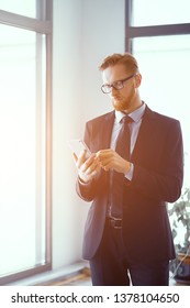 Businessman In Suit And Tie Standing At Window In Sunlight. Typing Message To Bussiness Partner. Business Concept.