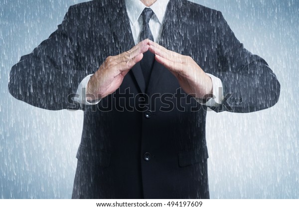 Businessman in suit with two\
hands in position to protect in rainy weather day. It indicates\
many aspects such as car insurance coverage, support, assurance,\
reliability.