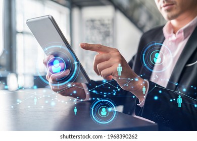 Businessman in suit using tablet device to check new candidates for international business consulting. HR, social media icons over modern panoramic office background.