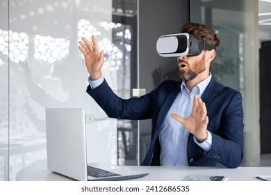Businessman in suit using VR headset at office desk, experiencing virtual reality technology with expressive hand gestures.