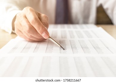 Businessman at table reading learning document report tables of digits and pointing at document with a silver pen. man in white shirt and blue tie. finance and accounting concept. selective focus