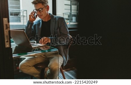 Businessman talking on mobile phone and looking at his laptop. Man sitting at his office desk working on laptop and discussing on phone.