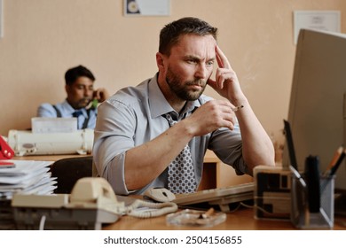 Businessman thinking while sitting at desk in office, wearing formal attire, with coworker in background using phone, surrounded by office supplies, showing thoughtful expression - Powered by Shutterstock