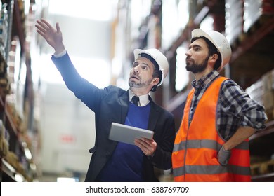 Businessman with touchpad pointing at upper shelf with goods while talking to worker