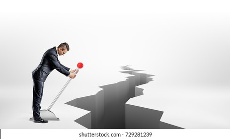 A Businessman Turns A Large Metal Lever While Standing Beside A Long Earthquake Rift On White Background. Change Circumstances. Use All You Can Get. Hostile Business Environment.