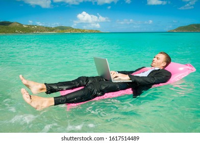 Businessman Typing On Laptop Floating On A Pink Sun Lounger In Bright Tropical Waters