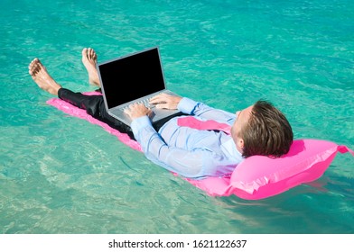 Businessman Typing On Laptop Floating On A Pink Sun Lounger In Bright Tropical Waters