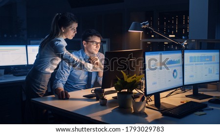 Businessman Uses Desktop Computer, His Female Project Manager Explains Specific Tasks, Account Handling and Strategic Moves. Professional People Late at Night in Big Corporate Office