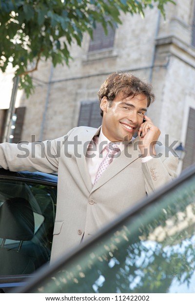 Businessman using a cell phone to make\
a phone call while standing some cars in the city,\
smiling.