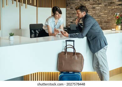 Businessman Using Digital Tablet To Checking In At Hotel.
Businessman With Luggage Standing At Hotel Reception And Filling In Registration Forms On Digital Tablet With Assistance Of Female Concierge.