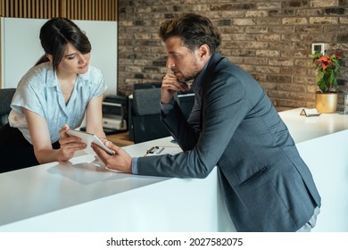 Businessman Using Digital Tablet For Checking In At Hotel.

Smiling Female Concierge Standing At Hotel Reception  And Showing Available Rooms On Digital Tablet To The New Guest.