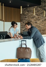 Businessman Using Digital Tablet To Checking In At Hotel. 
Businessman With Luggage Standing At Hotel Reception And Filling In Registration Forms On Digital Tablet With Assistance Of Female Concierge.