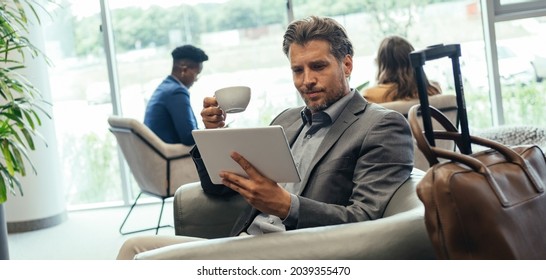 Businessman Using Digital Tablet And Drinking Coffee While Waiting For His Flight In The Airport VIP Lounge. 
Smiling Business Man Sitting In Armchair And Working On Tablet While Holding Cup Of Tea.