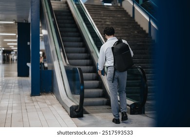 Businessman using an escalator in a modern indoor public space, wearing a backpack and holding a notebook. Represents urban commuting, business travel, and professional lifestyle. - Powered by Shutterstock