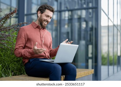 Businessman using laptop for digital meeting outdoors. Smiling in casual attire with earphones, seated on bench with modern building background. Depicts communication, technology, success. - Powered by Shutterstock