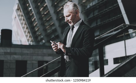 Businessman Is Using A Mobile Phone. Man Is Watching On Cellphone. Businessman Is Old Smiling Man. Man Wearing In Black Suit. Man Is Walking On Staircase Of Skyscraper. Sunny Daytime.