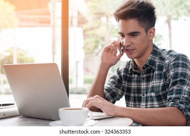 Businessman using mobile phone while looking at laptop on wooden table in coffee shop