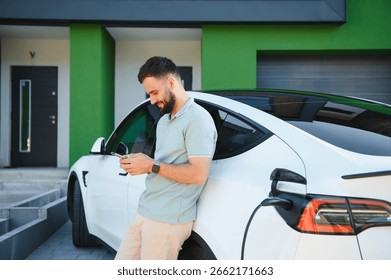 Businessman using phone while his electric car is charging at his house
