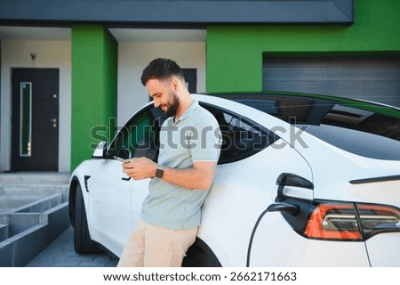 Businessman using phone while his electric car is charging at his house