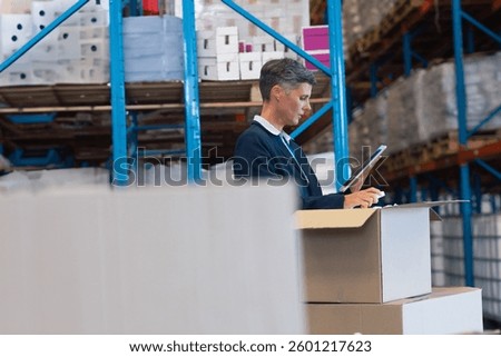 Businessman using tablet checking inventory in large warehouse with shelves, copy space. Logistics, supply chain, industrial, storage, management, technology
