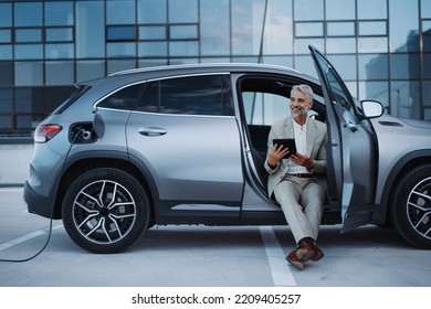Businessman using tablet while charging car at electric vehicle charging station, close-up. - Shutterstock ID 2209405257