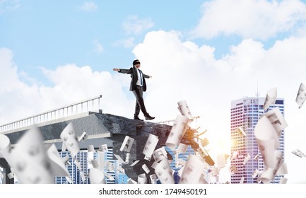 Businessman walking blindfolded among flying documents on concrete bridge with huge gap as symbol of hidden threats and risks. Cityscape and sunlight on background. 3D rendering.