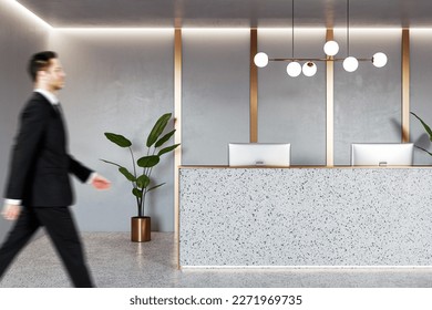 Businessman walking by stylish reception desk with modern computers in office area with green plant and golden decoration wall background