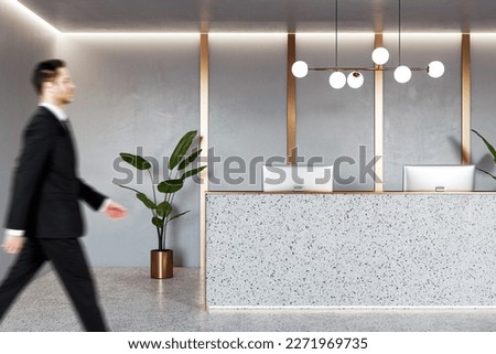 Businessman walking by stylish reception desk with modern computers in office area with green plant and golden decoration wall background