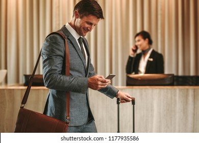 Businessman Walking In Hotel Lobby With Suitcase And Using His Smart Phone. Male Business Traveler In Hotel Hallway With Cellphone And Luggage.