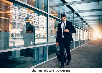 Businessman walking with luggage and using mobile phone at airport. Young man on business trip text messaging from his cell phone. - Powered by Shutterstock