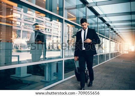 Businessman walking with luggage and using mobile phone at airport. Young man on business trip text messaging from his cell phone.