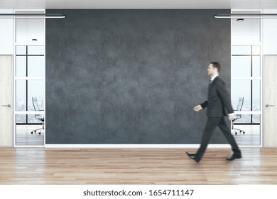 Businessman Walking In Modern Empty Office Hall With Copy Space On Black Wall And Wooden Floor.