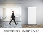 Businessman walking in modern glass office interior with empty mock up banner on wall, wooden flooring, furniture, window with city view and other objects