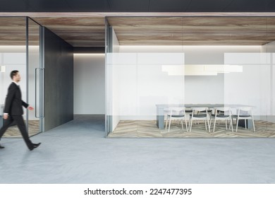 Businessman walking on concrete floor by stylish conference rooms with minimalistic style furniture on wooden parquet floor behind glass partitions in spacious office hall