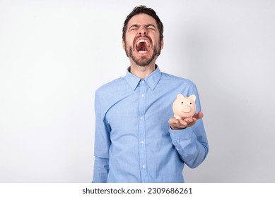 businessman wearing blue shirt holding a piggy bank  over white background angry and mad screaming frustrated and furious, shouting with anger. Rage and aggressive concept. - Shutterstock ID 2309686261