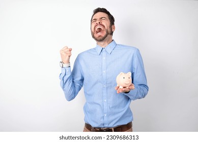 businessman wearing blue shirt holding a piggy bank  over white background celebrating surprised and amazed for success with arms raised and eyes closed. Winner concept. - Shutterstock ID 2309686313