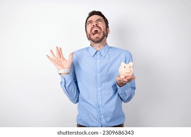 businessman wearing blue shirt holding a piggy bank  over white background crazy and mad shouting and yelling with aggressive expression and arms raised. Frustration concept. - Shutterstock ID 2309686343