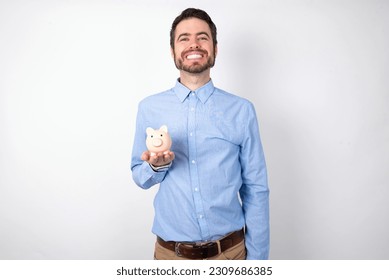 businessman wearing blue shirt holding a piggy bank  over white background with a happy and cool smile on face. Lucky person.