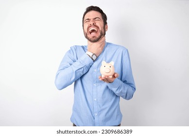 businessman wearing blue shirt holding a piggy bank  over white background shouting suffocate because painful strangle. Health problem. Asphyxiate and suicide concept. - Shutterstock ID 2309686389