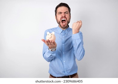 businessman wearing blue shirt holding a piggy bank  over white background angry and mad raising fist frustrated and furious while shouting with anger. Rage and aggressive concept. - Shutterstock ID 2309686405