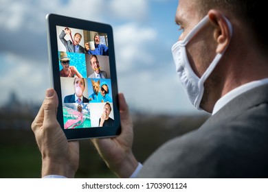 Businessman Wearing Coronavirus Face Mask Holding His Remote-work Tablet Showing A Grid Of Self-isolating Attendees To A Web Video Conference