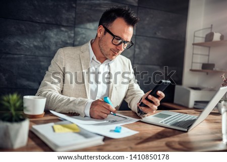 Businessman wearing linen suit and eyeglasses sitting at his desk using smart phone and highlighting text with blue marker in his office