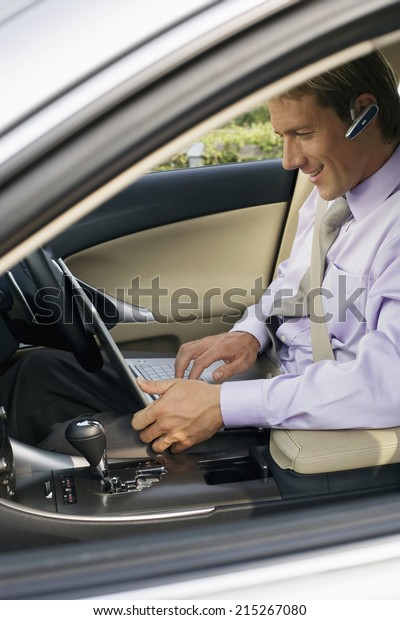 Businessman wearing mobile phone\
hands-free device, sitting in car, using laptop, side\
view