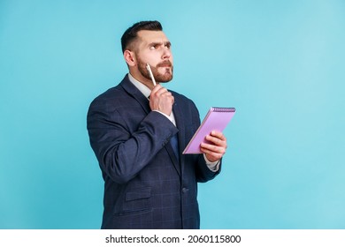 Businessman wearing official style suit pondering idea to write into notebook, taking notes in paper, thinking over business plan, to-do list. Indoor studio shot isolated on blue background.